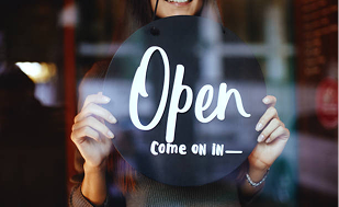 A person is holding a round black sign with the word “Open” and the phrase “Come On In” written in white letters. The sign is positioned in front of a glass door or window, indicating that the business is welcoming customers. The background shows a warm indoor setting with soft lighting.
