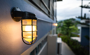 A close-up view of a warm glowing outdoor wall light fixture mounted on the exterior of a modern building. The fixture has a protective metal cage design and emits a soft yellow light. The background shows a blurred outdoor setting with greenery and architectural elements, suggesting evening or early night lighting.
