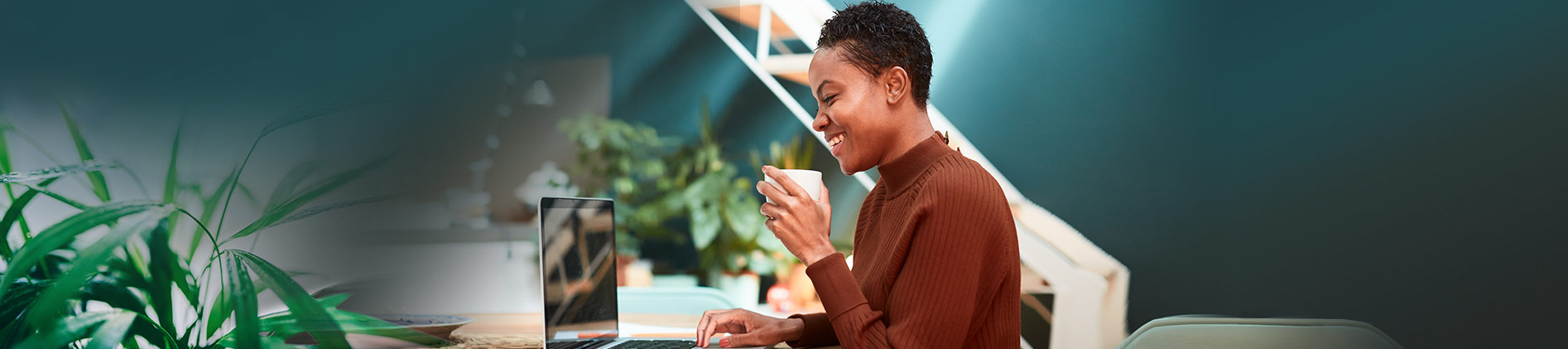 Woman smiling and sitting at a desk while drinking coffee and typing on her laptop